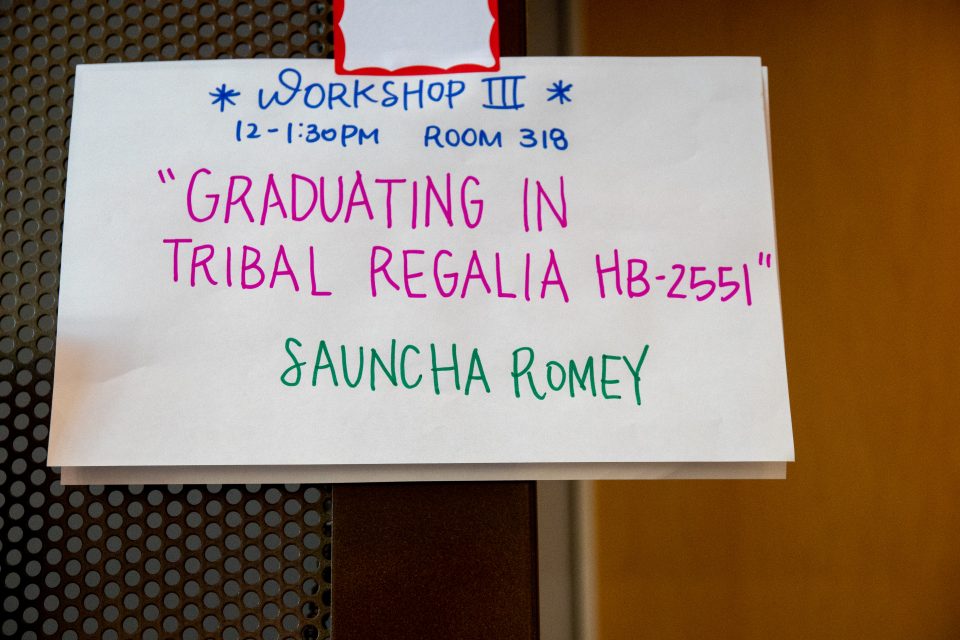 A sign that says "Workshop 3 12-1:30 PM Room 318. Graduating in Tribal Regalia HB 2551" Sauncha Romey