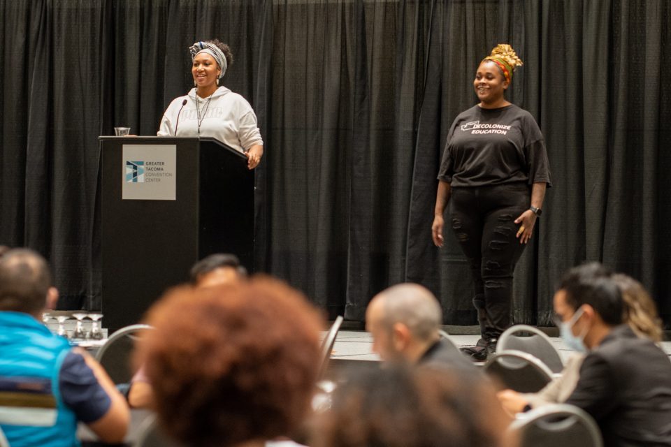 Two people standing on stage with some standing behind a podium