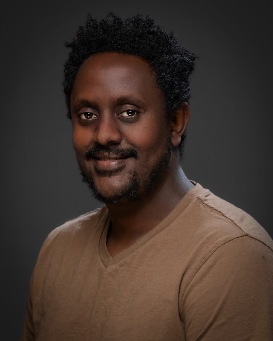 Abas, a Black man in his early 30s with black hair and a well-groomed beard, wearing a regular shirt, smiling warmly at the camera.