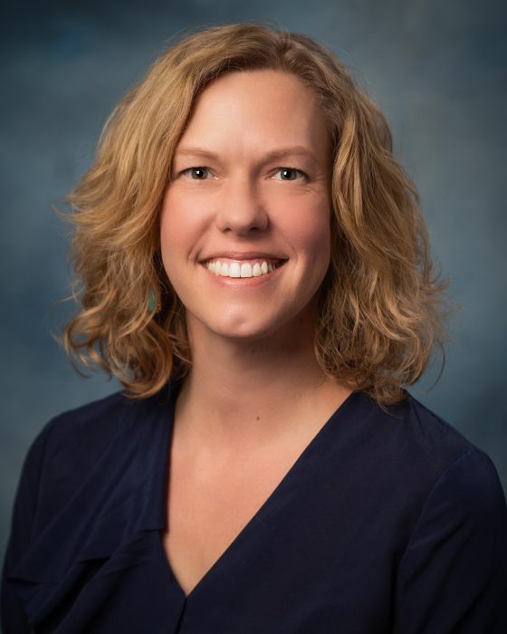 A white woman in her early 50s with chin length blonde wavy hair, wearing a navy blue blouse smiling at the camera