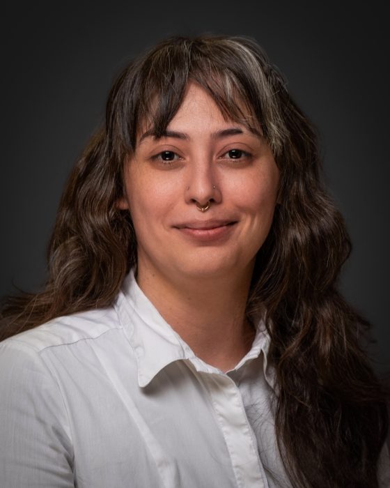 Jeanina Messerly, a Chicano person in their early 30's with long wavy brown hair wearing a white button up shirt.