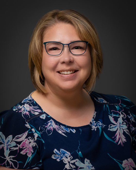 Jennifer Youngblood, a white-presenting woman in her late 40s, with straight blonde hair and glasses, wearing a blue shirt with flowers, smiling at the camera.