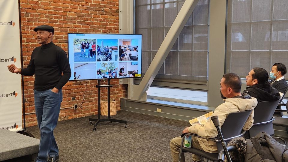 A man standing at the from of a room talking to people sitting in chairs looking at a TV screen