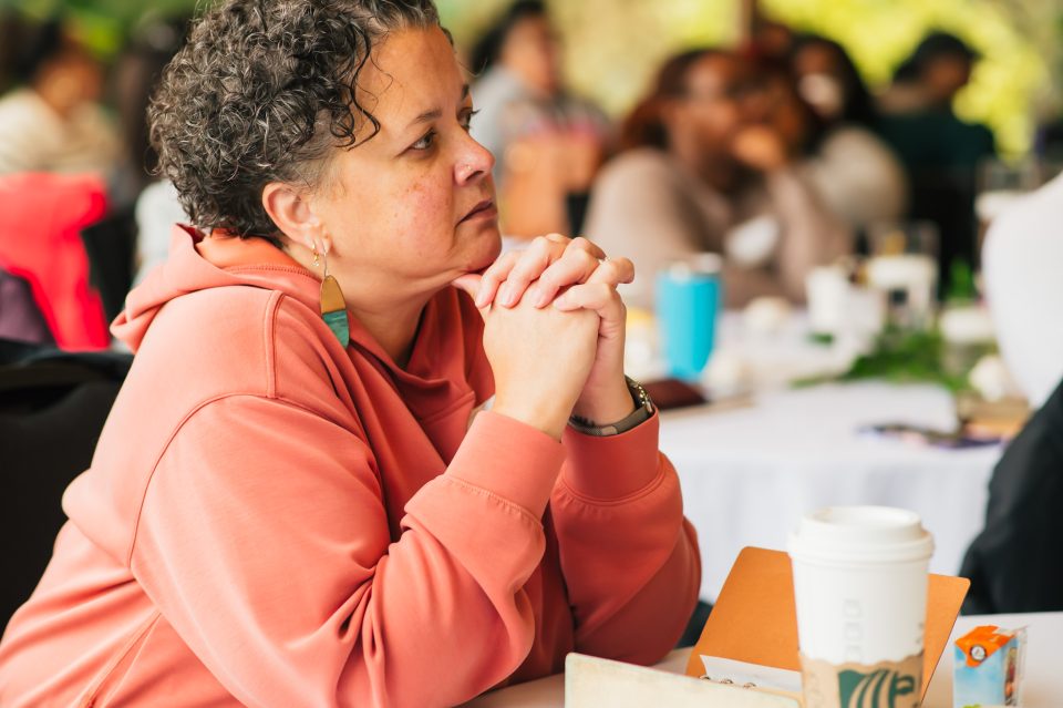Carlina Brown Banks, a person with short curly hair leaning againsta table in a coral colored sweatshirt