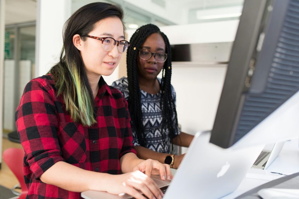Woman Wearing Red and Black Checkered Blouse Using Macbook with another woman looking next to her